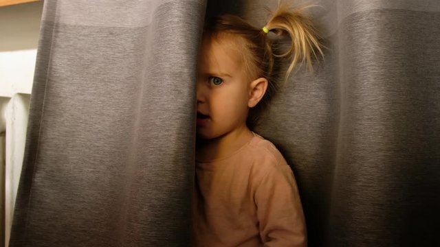 Scared little girl in casual wear standing behind curtain and looking at camera with suspicion