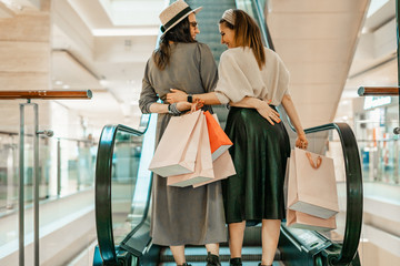 Two girls on an escalator in a shopping center. In the hands of paper bags. Shopping, leisure and entertainment.