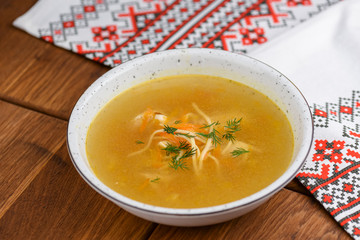 Chicken soup with noodles and vegetables served in white bowl over rustic wooden table with traditional tablecloth.