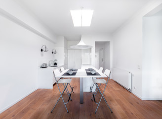New stylish bright kitchen with white cabinets. Spacious modern interior with wooden floor, white table, industrial chairs and big ceiling window.