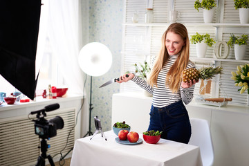 Food blogger cooking fresh vegan salad of fruits in kitchen studio, filming tutorial on camera for video channel. Female influencer holds apple, pineapple and talks about healthy eating. Fructorianism