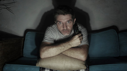 young attractive concentrated and scared man holding TV remote watching horror movie or suspense...