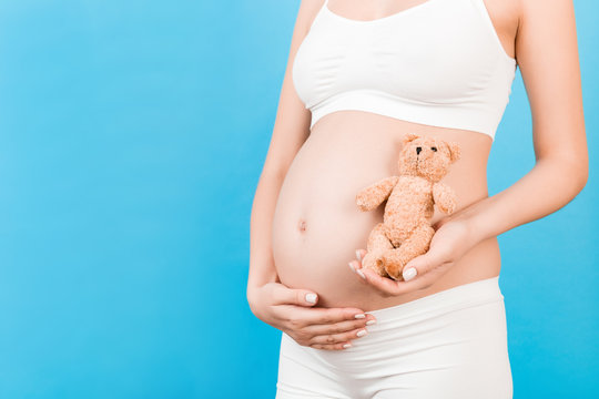 Cropped image of happy pregnant woman in white underwear holding teddy bear against her belly at blue background. Child expecting. Copy space