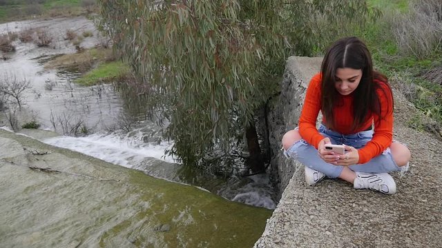Female Model Updating Social Media On Phone In Park Next To River In Slow Motion