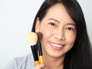 Asian woman has dark brown long hair, is makeup artist, holding brush and make up cheek bone aspink nude tone. Lady is smiling, happy and cheerful to be beautiful stylelist concept.  Gray t-shirt.