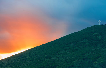 Sunset sky over a green mountain with a white cross monument on top