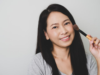 Asian adulth woman is smiling and  happy. She use brush on cheekbone to makeup as nude warm bright matte tone color.  Look casual clothes.  Matte tone.