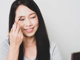 Asian adulth woman is smiling and  happy. She use brush on cheekbone to makeup as nude warm bright matte tone color.  Look casual clothes. Matt Tone
