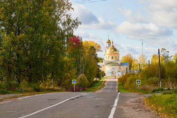 Asphalt road leading to the village with a beautiful white stone Orthodox Church with a yellow dome among nature and green trees. Natural landscape with greenery on a summer, autumn or spring day