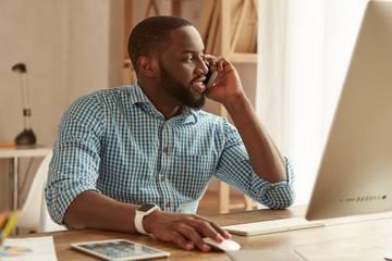 Good talk. Happy afro american businessman in shirt talking by phone with client and smiling while working on the computer at his working place at home. Freelance