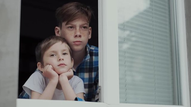 Two brothers of 5 and 13 years of age sadly watch the street from their window. Concept: Children in quarantine are deprived of walks in the fresh air.