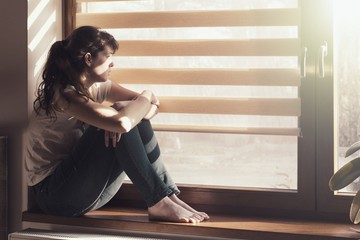 Sad young woman sitting on the window at home isolated, watching out. Coronavirus quarantine...