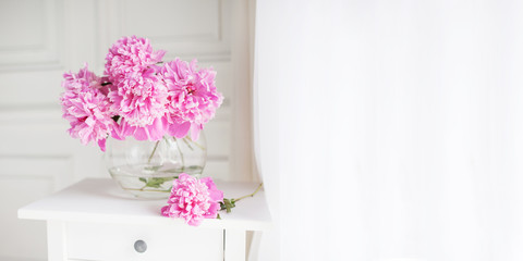 Pink peonies in glass vase. Flowers on a white table near the window.  Beautiful peony flower for catalog or online store. Floral shop and delivery concept. Banner. Copy space