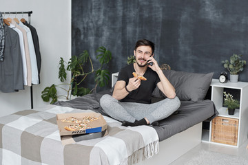 Obraz premium Cheerful handsome young bearded man sitting on bed and chatting by phone while eating pizza at home