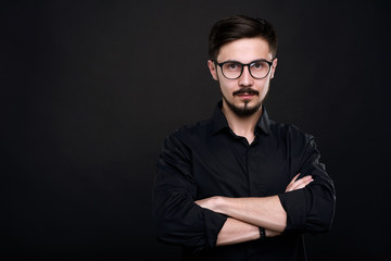 Obraz premium Portrait of serious confident young entrepreneur with mustache and beard standing with crossed arms against black background