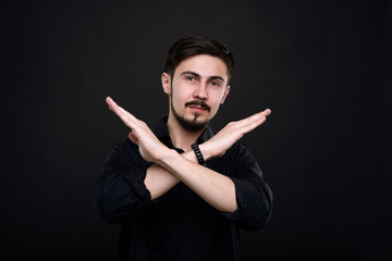 Portrait of young bearded man standing against black background and crossing arms while making stop gesture