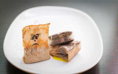On a white plate are used tea bags, a used tea bag with a drawn sad face.