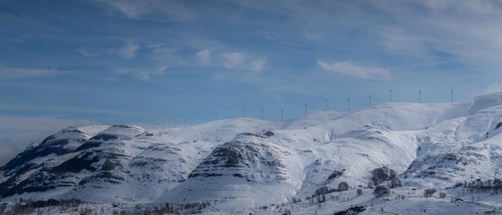 Panorama shot of windmills in a snowed mountain with blue sky.