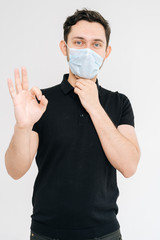 Caucasian young man in medical mask and black t-shirt showing ok sign on white back on isolated white background. Prevention of virus infection. Concept of Coronavirus COVID-19 Pandemic.