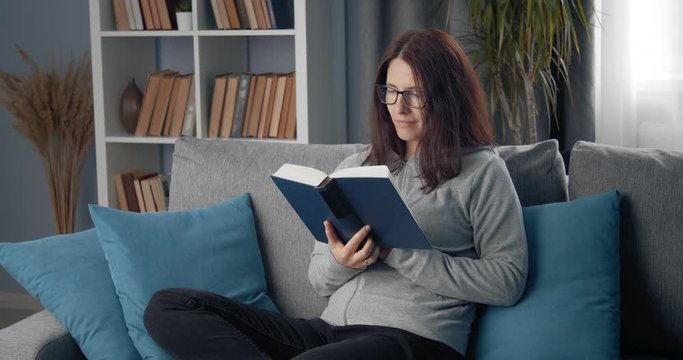 Cheerful lady in eyeglasses and domestic clothing sitting on couch with crossed legs and reading book. Concept of comfort, leisure and literature.