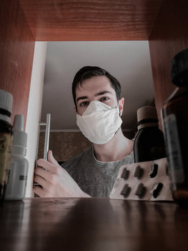 European Man In A Medical Mask Looks At Medication In Medicine Cabinet. View From Medicine Cabinet At Home. Coronavirus Concept.