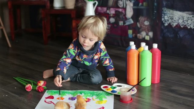 Sweet toddler blond child, boy, painting with colors, little chicks walking around him, making funny prints on his paper