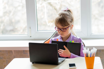 A little schoolgirl sits at a desk and studies on a laptop. The concept of distance learning.