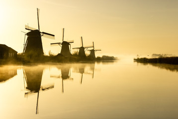 Historic dutch windmills along a wide canal at Kinderdijk in a polder wetland on a foggy morning in...
