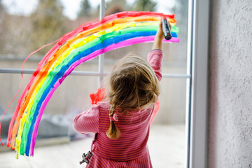 Adoralbe little toddler girl with rainbow painted with colorful window color during pandemic...