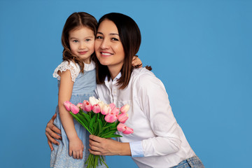 portrait of a happy family on a blue background. mother and daughter with a white teeth smile look at the frame and hold a bouquet of pink tulips in their hands