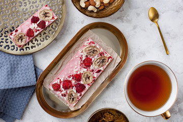 handmade pink chocolate bar with banana, raspberry and puffed rice lying on the table near a cup of tea