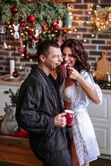 People in dressing gowns in the kitchen, which is decorated for Christmas. A girl treats a guy to a Cup of cocoa