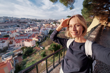 Fototapeta na wymiar Traveling by Portugal. Young traveling woman taking selfie in old town Lisbon with view on red tiled roofs, ancient architecture.