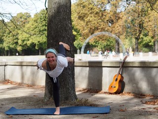 Girl doing a yoga pose outside in a park infront of a tree with a guitar leaning next to a wall.