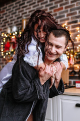 A guy and a girl in dressing gowns in the kitchen, which is decorated for the celebration of Christmas and New year. The girl climbed on the guy's back