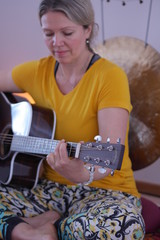 Close up of a woman in a yellow T-Shirt playing the guitar in a yoga class with a gong in...