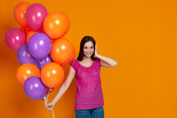 Fototapeta na wymiar Smiling delighted girl in pink t-shirt posing with bright colorful air balloons isolated over orange background. Beautiful happy young woman on a birthday party. space for text