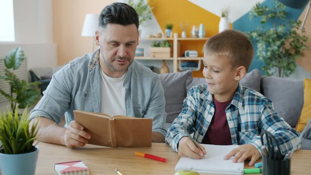 Creative child is drawing pictures in notebook while loving father is reading book sitting at desk together at home. People, lifestyle and culture concept.
