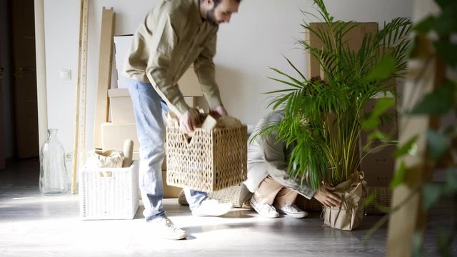 joyful young family of happy guy and girl wearing casual clothes carries boxes with different belongings into large room of new light apartment