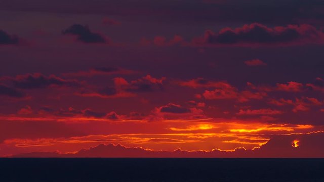 An almost still and beautiful summer sunset / sundown over a calm sea, with orange / pink / red light throughout the sunlit cloudy sky. Taken off the coast of Cornwall, England, UK.