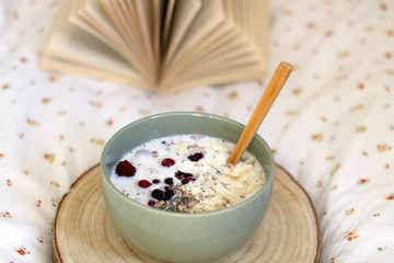 Breakfast bowl with milk, oats, chia seeds and berries, served on a bed with floral sheets. Open book in the background. Selective focus.