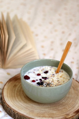 Breakfast bowl with milk, oats, chia seeds and berries, served on a bed with floral sheets. Open book in the background. Selective focus.