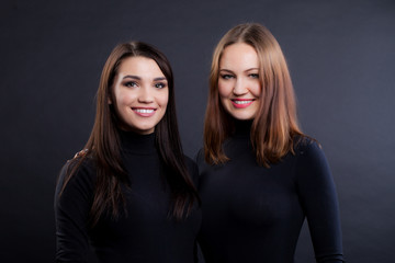 Two young girlfriends are standing together. Both are in black clothes and are looking at the camera. Black background