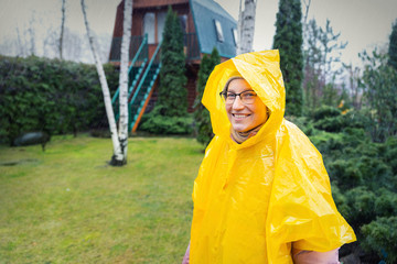 Portrait of beautiful young adult caucasian woman under bright yellow raincoat and wellies walking at garden during cold spring rainy day at home backyard. Carefree hipster concept. Bad weather