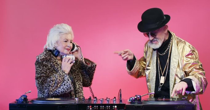 Stylish disco couple of retired DJs mixing music on audio equipment. Fanny retro style Caucasian grey-haired lady in headphones and senior man in golden jacket dancing over pink wall. Party concept