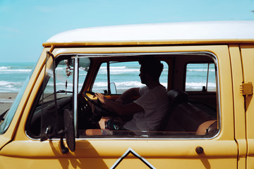A young man is sitting at his oldtimer van looking for good waves for surfing. Enjoying his...