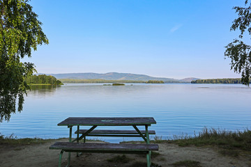 Lake shore and empty picnic bench. Summer landscape of a lake or river with calm water and a park furniture to relax near the lakeside. Beautiful and tranquility view of Sungul lake in Russia.