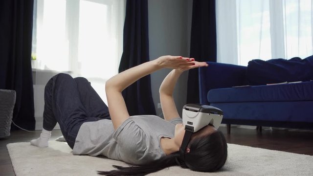 Young Asian woman in vr headset exploring virtual reality lying on carpet on floor at home