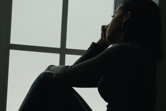 Profile Portrait Of A Frustrated African American Woman With Straight Black Hair In The Dark Staying Beside The Window And Close Her Face With Arms