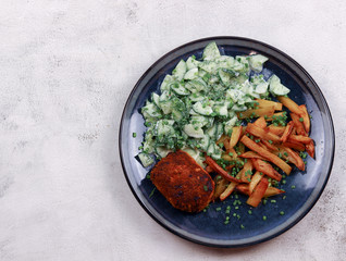 Fried potatoes with cucumber salad and chicken patty on a round plate on a light gray background....